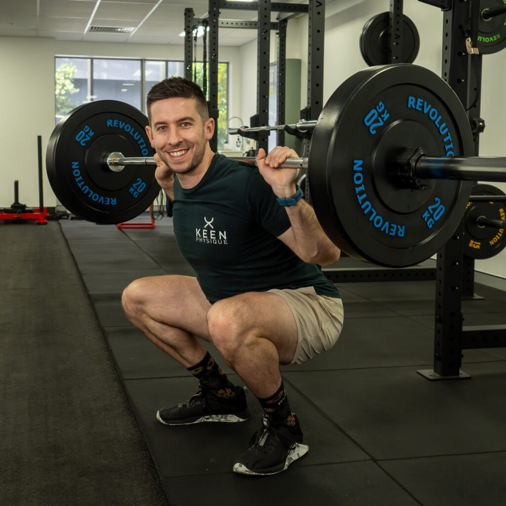 Podiatrist and personal trainer demonstrating a barbell squat while smiling at the camera.